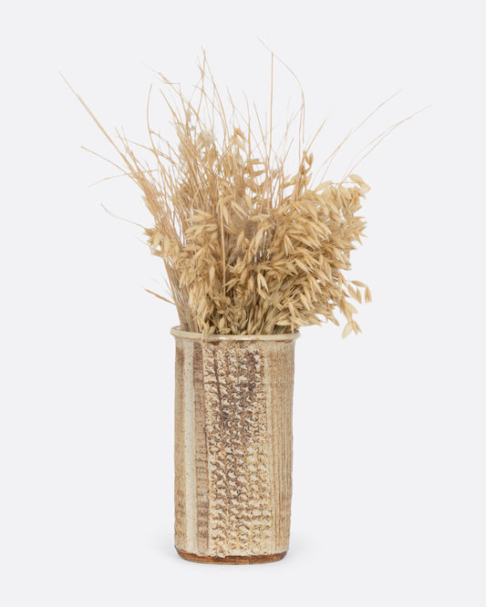 A tall textured tan vase. Shown with grasses in it from the front. 