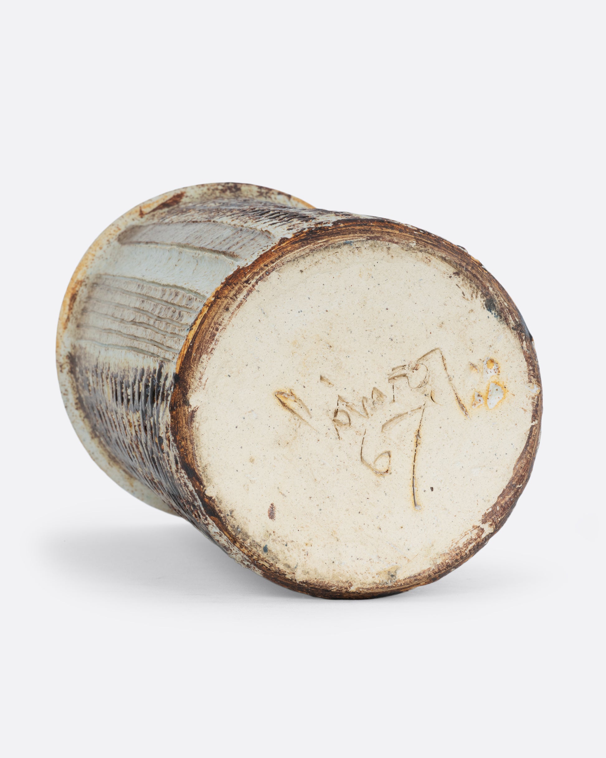 A tall textured tan vase with the artist signature and 67 on the bottom. Shown on its side from the bottom. 