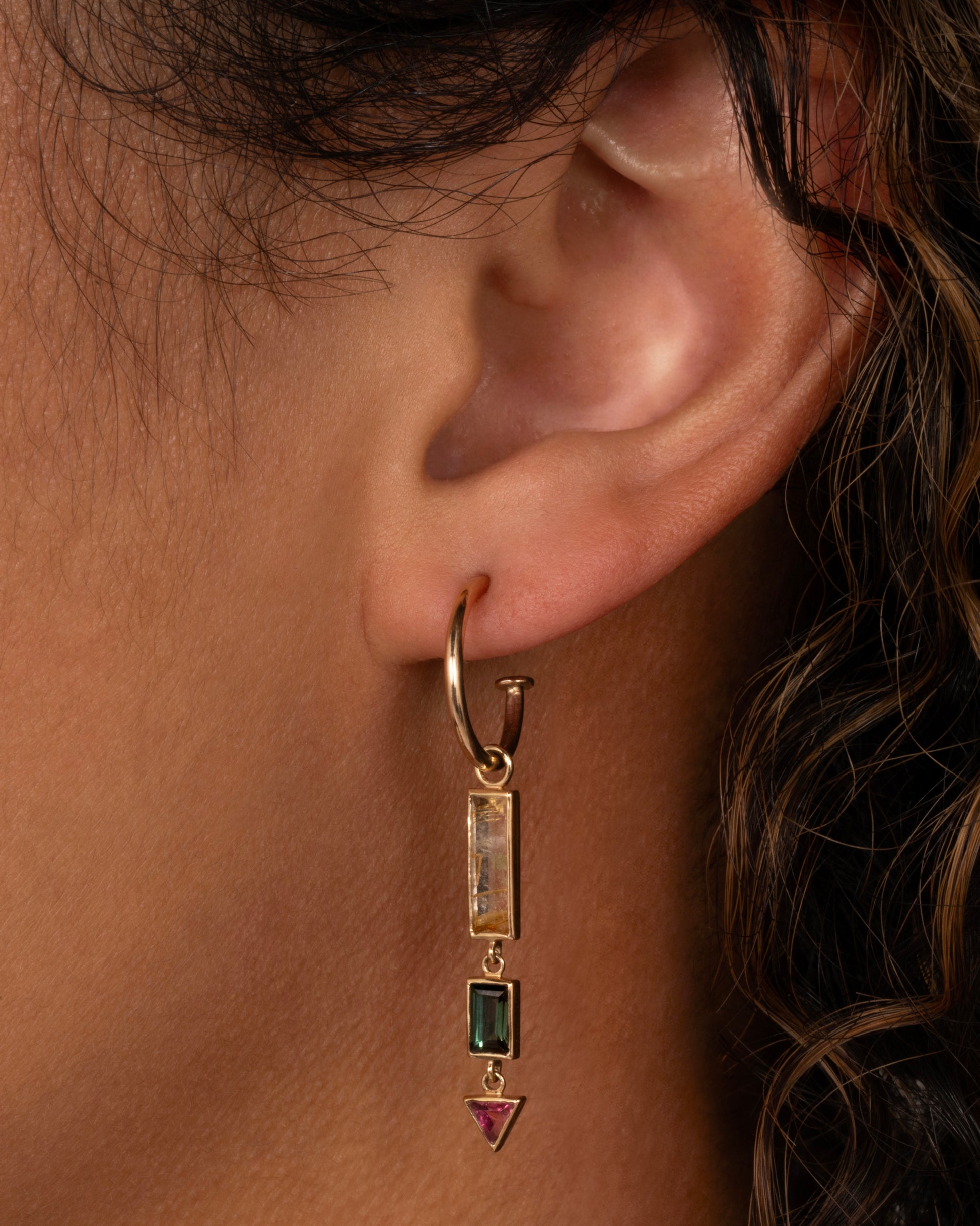 A yellow gold dangle with a rectangular rutilated quartz top, a small green rectangular tourmaline, and a pink tourmaline triangle. Shown on a yellow gold hoop from the front on a person.