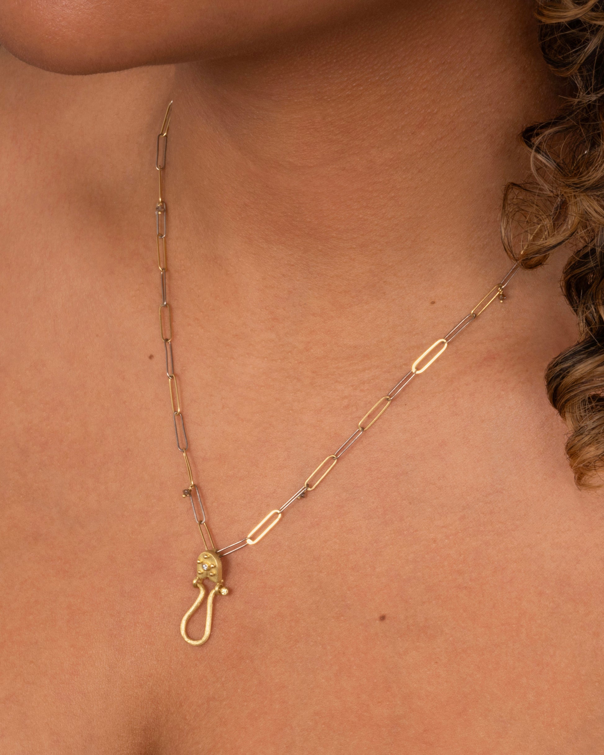 A yellow gold charm holder pendant with white diamonds at the top and at the end of the loop. Shown on a yellow gold paperclip chain on a person from the top front.