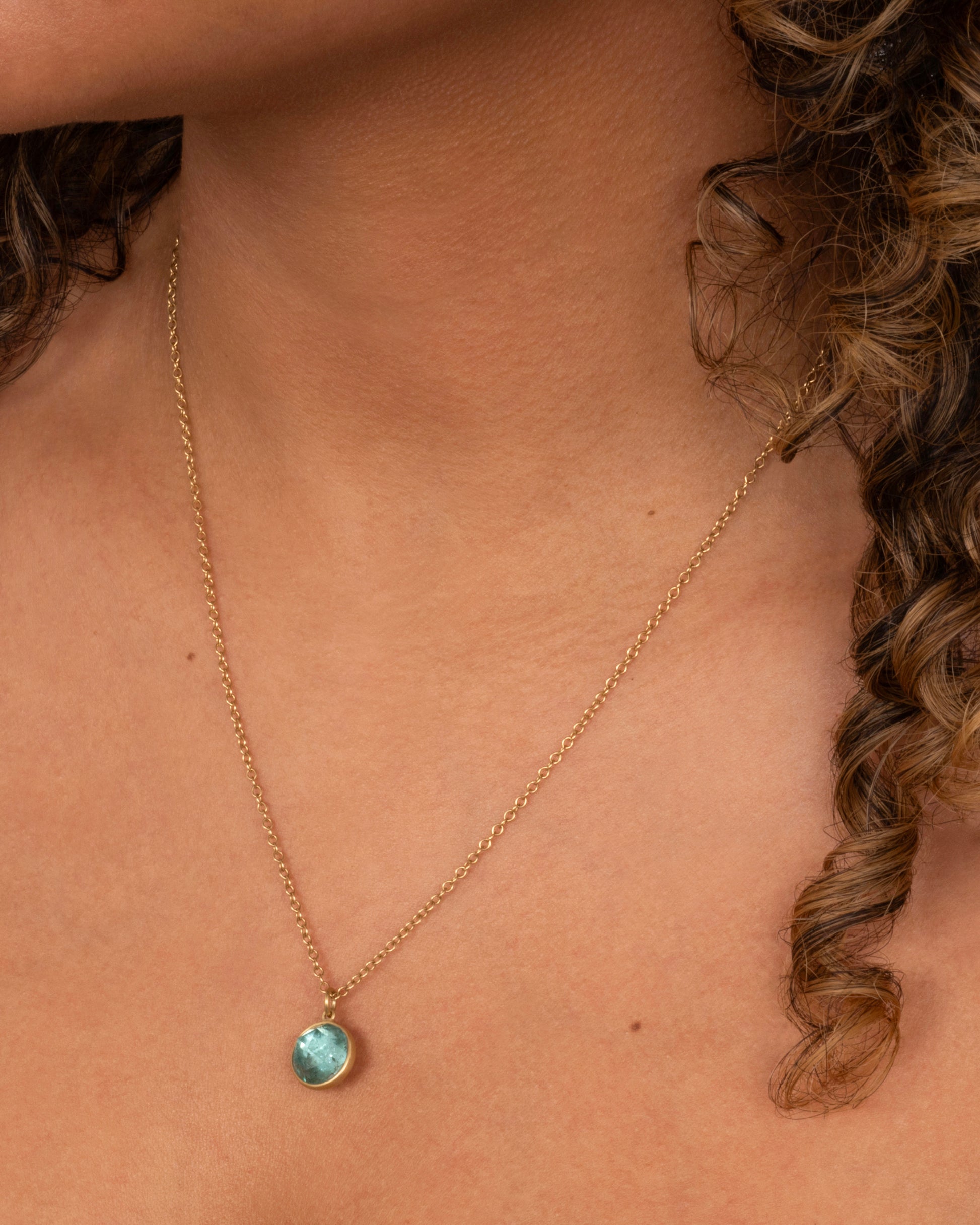 A yellow gold necklace with a circular light blue tourmaline pendant. Shown worn around the neck.