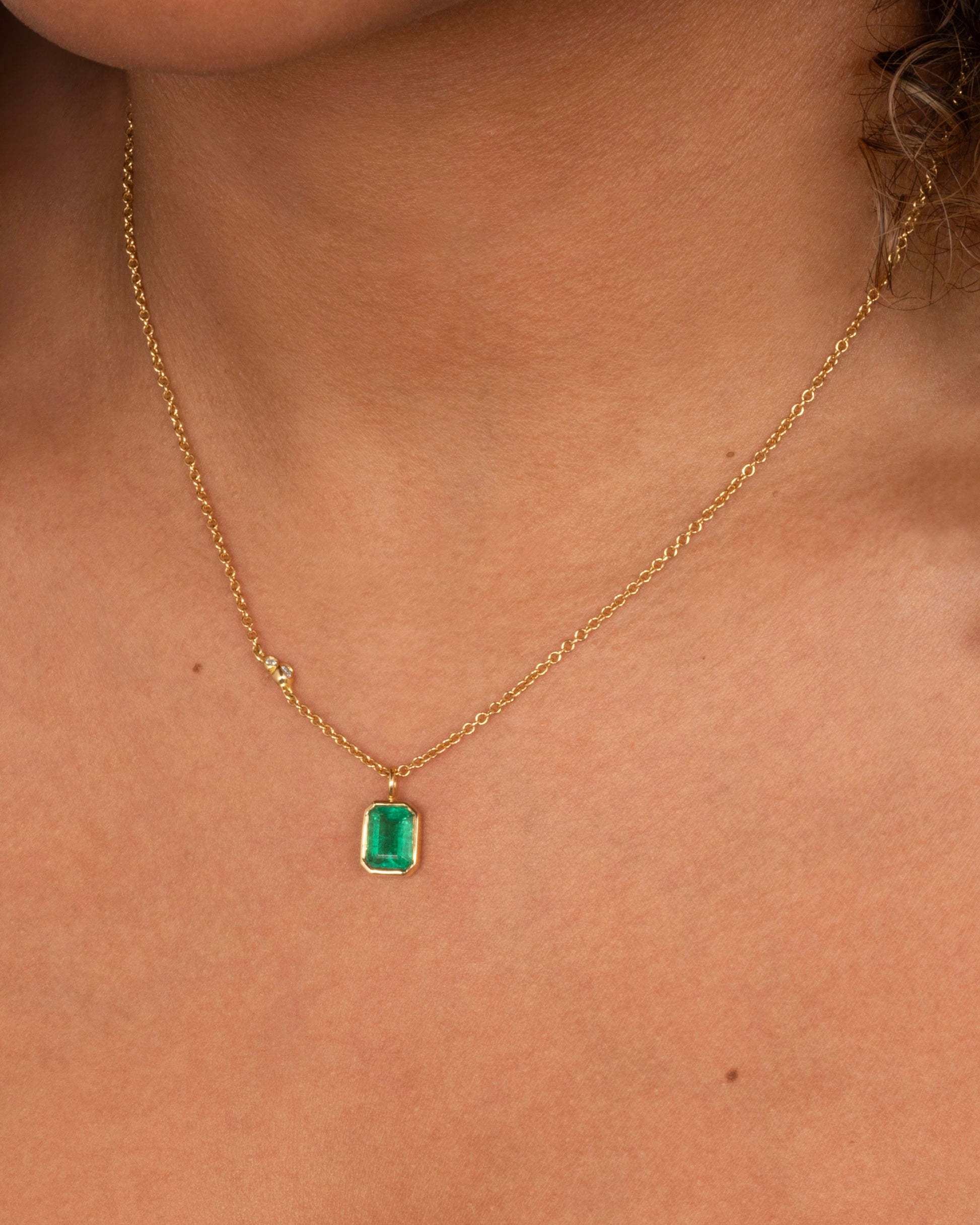 A yellow gold cable chain necklace with a bezel set Zambian emerald and diamond details on one side. Shown from the front on a person.