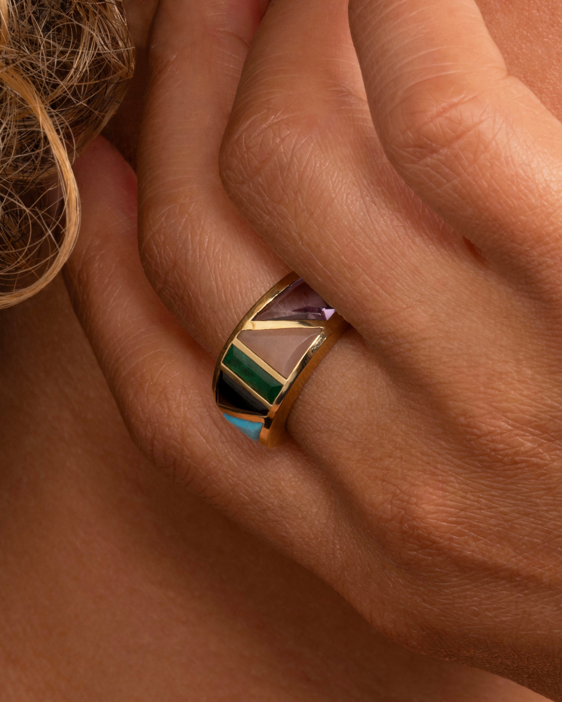 A yellow gold band with an array of gemstones in angular shapes. Shown worn on a hand.