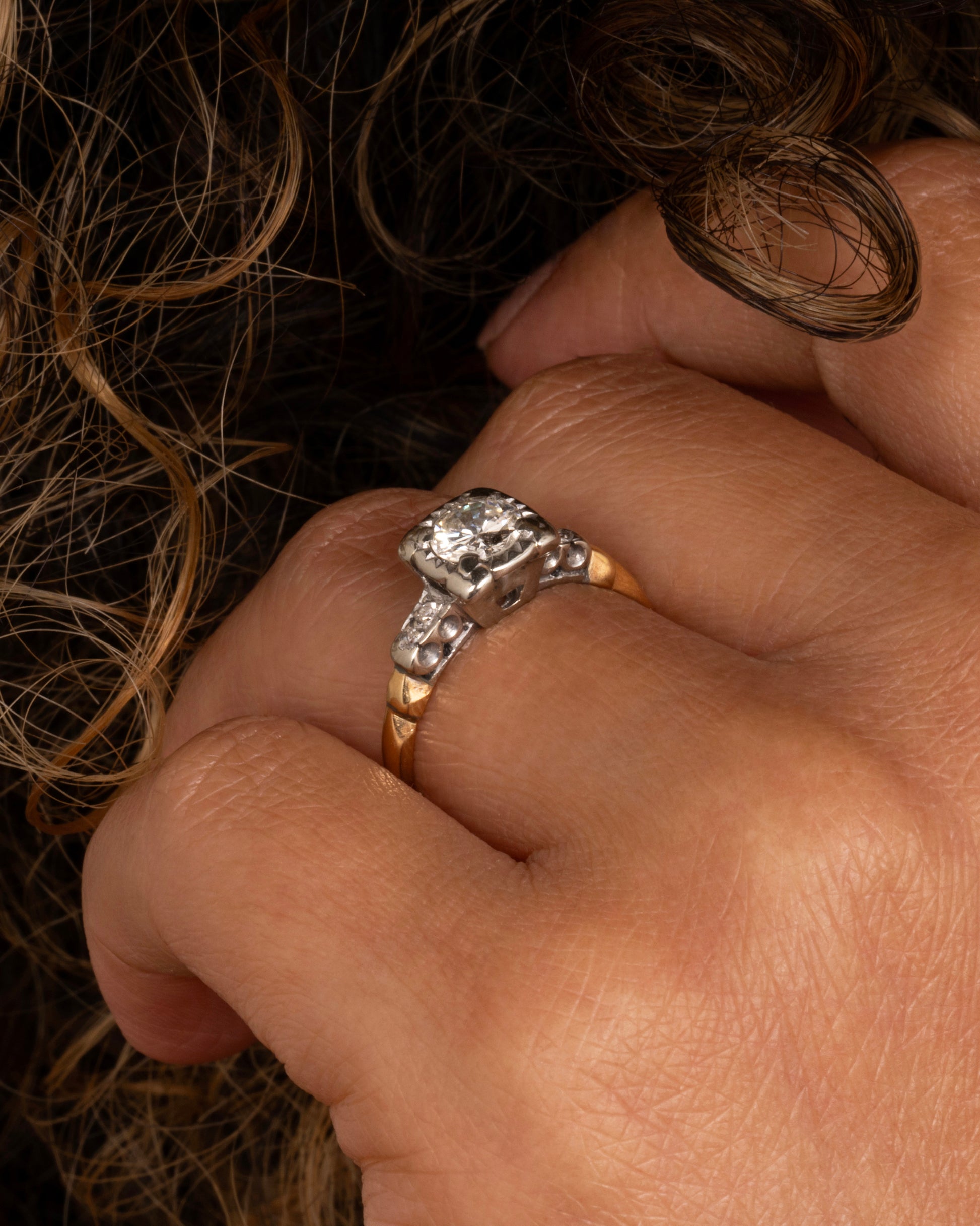 A yellow gold ring with a white gold setting and a round central diamond with four bead set diamonds on the shoulders. Shown worn on a hand.