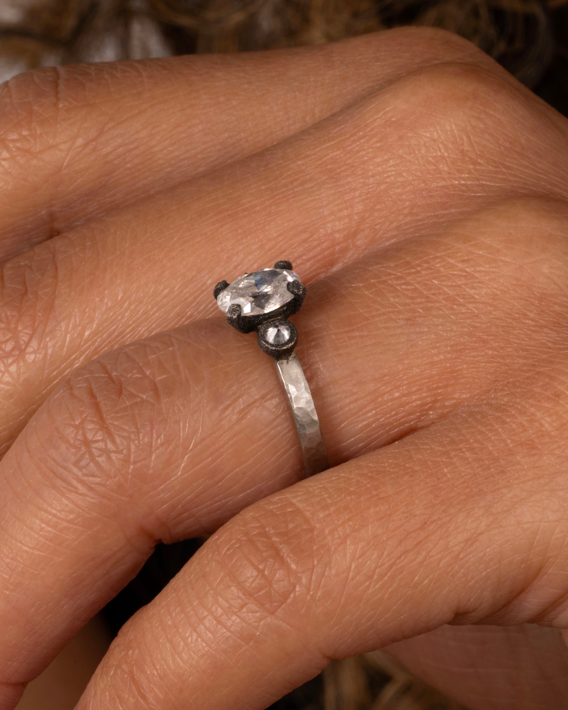 A hammered palladium band with three diamonds set in darkened white gold settings; one larger oval diamond flanked by two inverted round diamonds. shown on a finger.