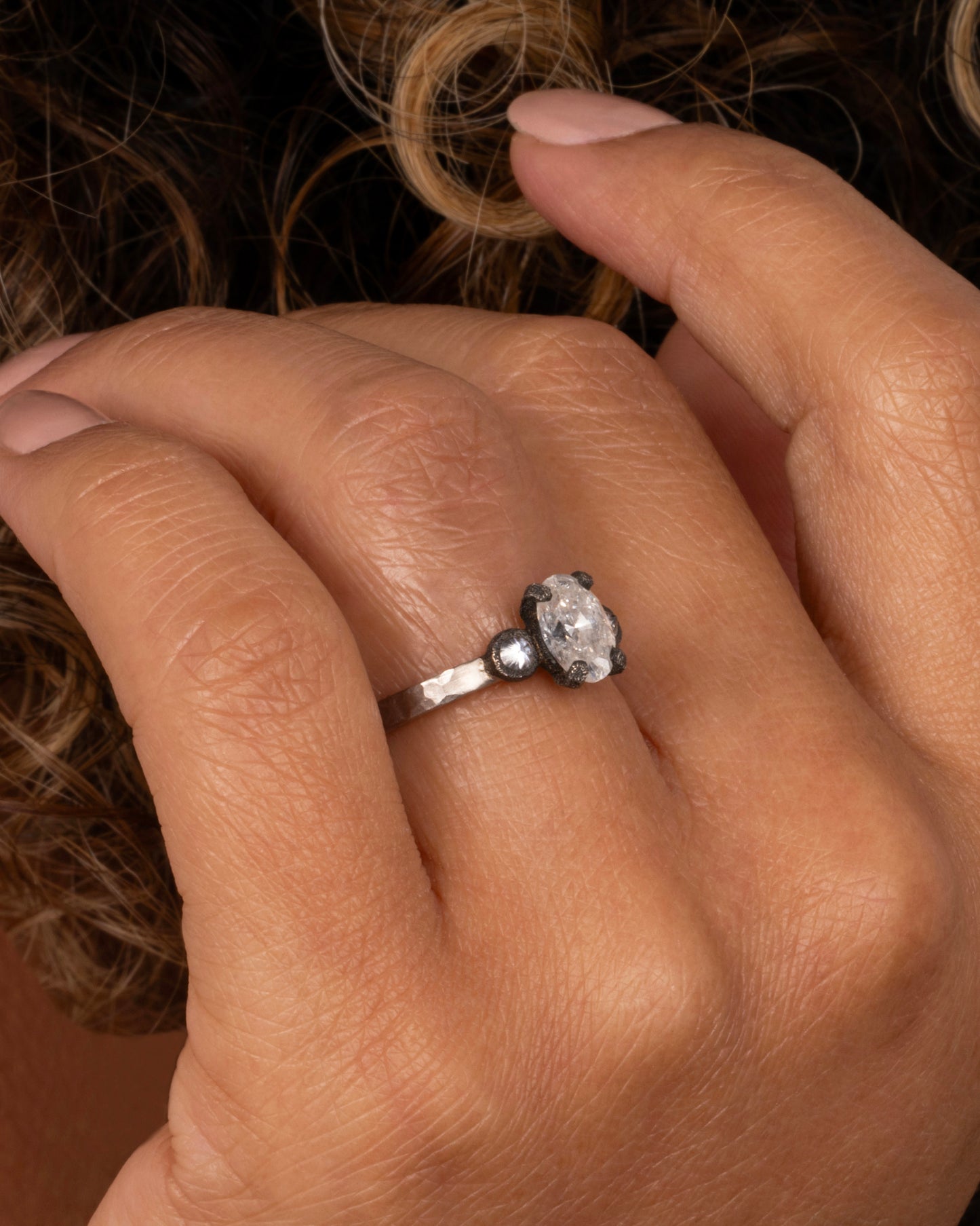 A hammered palladium band with three diamonds set in darkened white gold settings; one larger oval diamond flanked by two inverted round diamonds. shown on a finger.