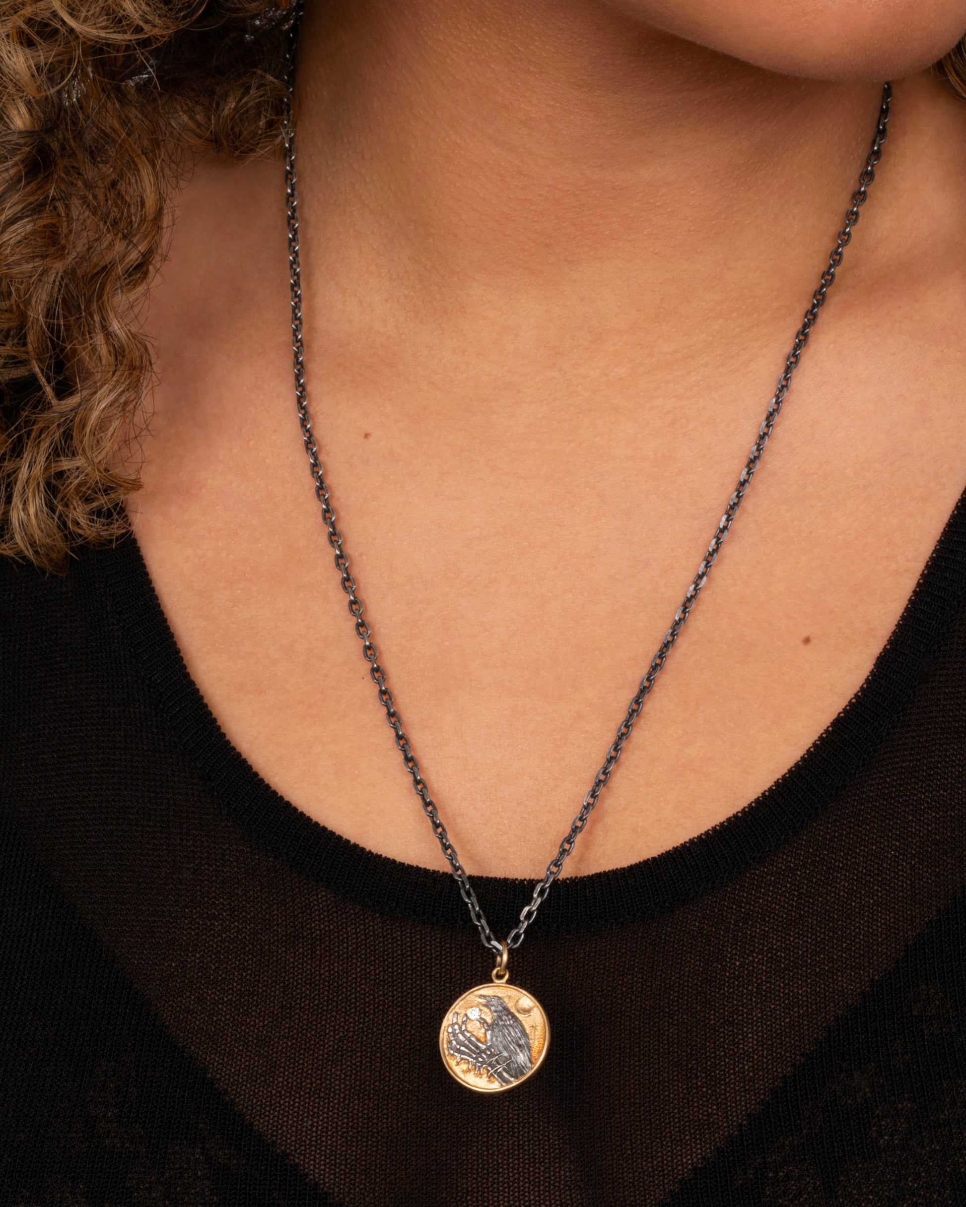 A yellow gold and silver pendant necklace with a skeleton hand holding a diamond and a crow. Shown worn around the neck.