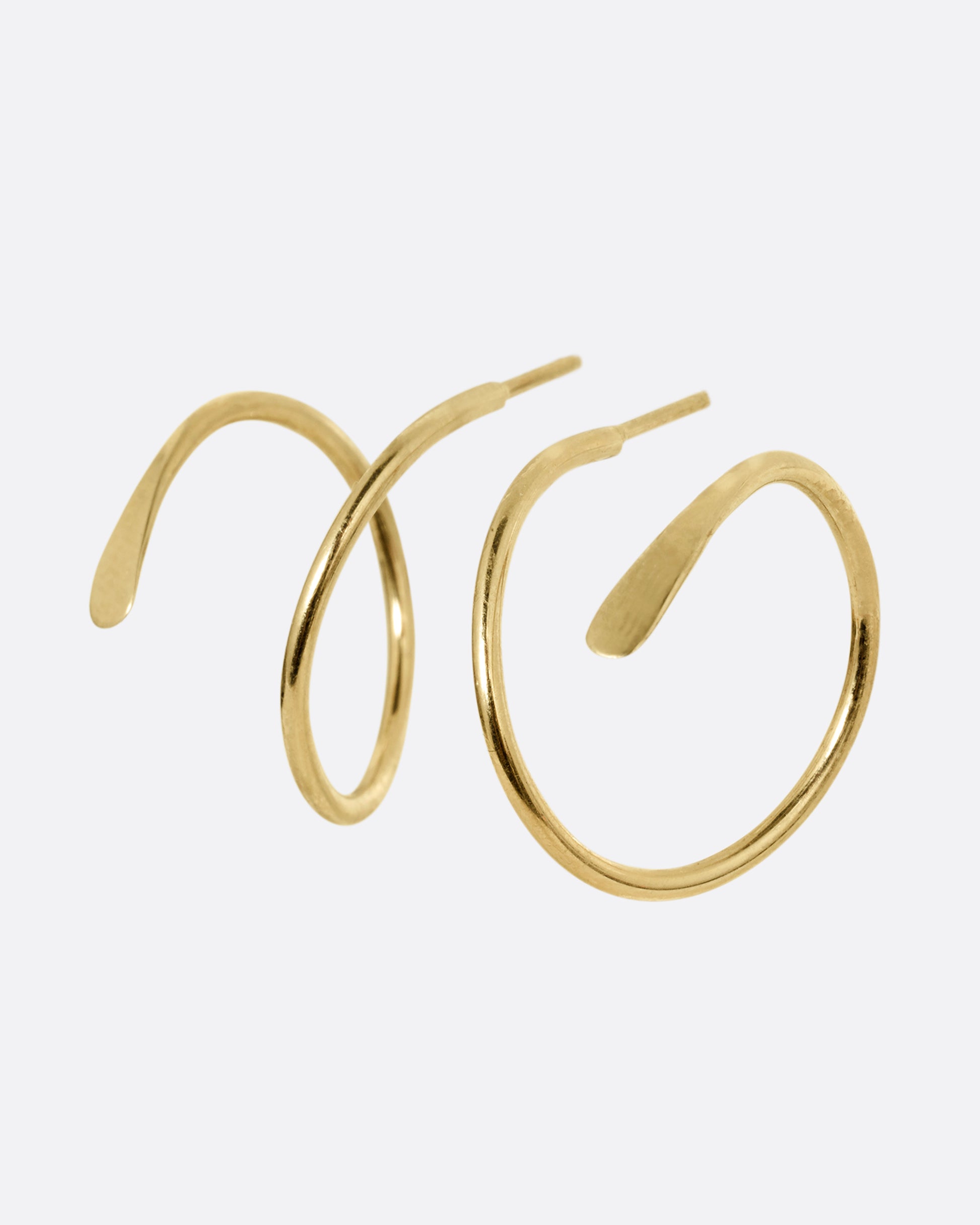 A pair of yellow gold earrings in a loop shape with one hammered flat end. Shown standing up from the front.