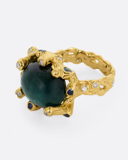 A yellow gold ring with a large indicolite tourmaline cabochon, surrounded by blue sapphires and diamonds, shown from the side.