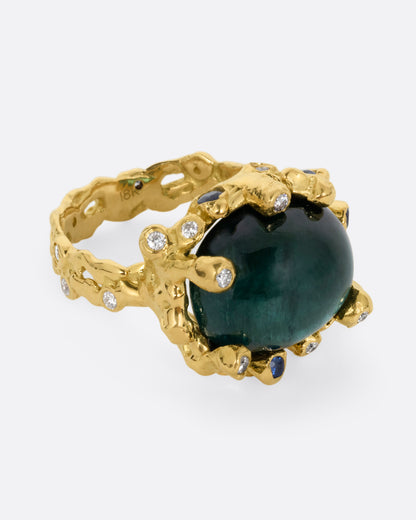 A yellow gold ring with a large indicolite tourmaline cabochon, surrounded by blue sapphires and diamonds, shown from the side.