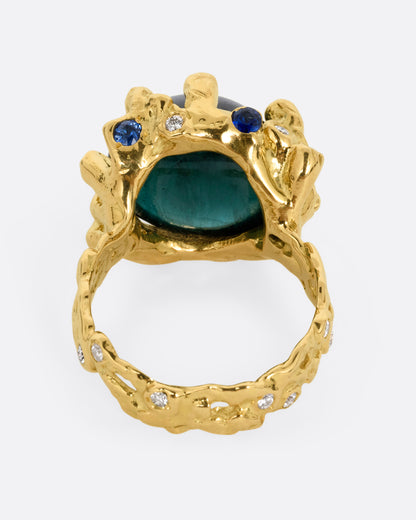 A yellow gold ring with a large indicolite tourmaline cabochon, surrounded by blue sapphires and diamonds, shown from the back.