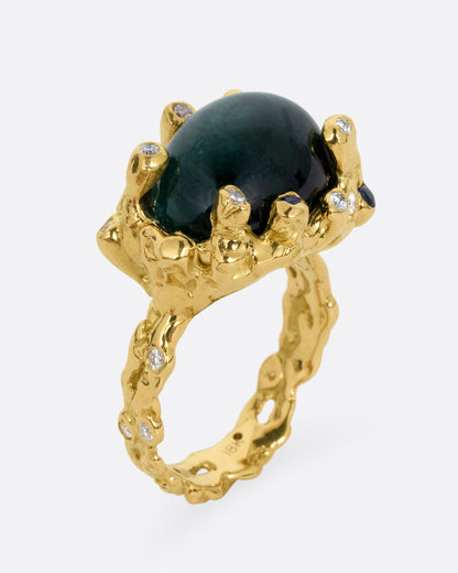 A yellow gold ring with a large indicolite tourmaline cabochon, surrounded by blue sapphires and diamonds, shown from the side standing up.