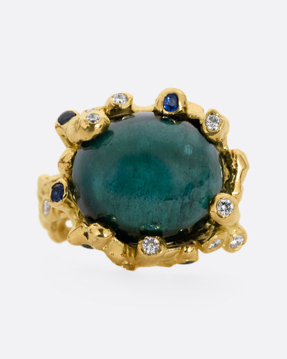 A yellow gold ring with a large indicolite tourmaline cabochon, surrounded by blue sapphires and diamonds, shown from the front. 