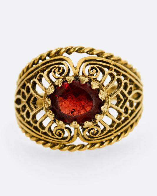 A yellow gold dome ring with an intricate web of filigree and delicate cutouts and a garnet at its center. Shown from the front.