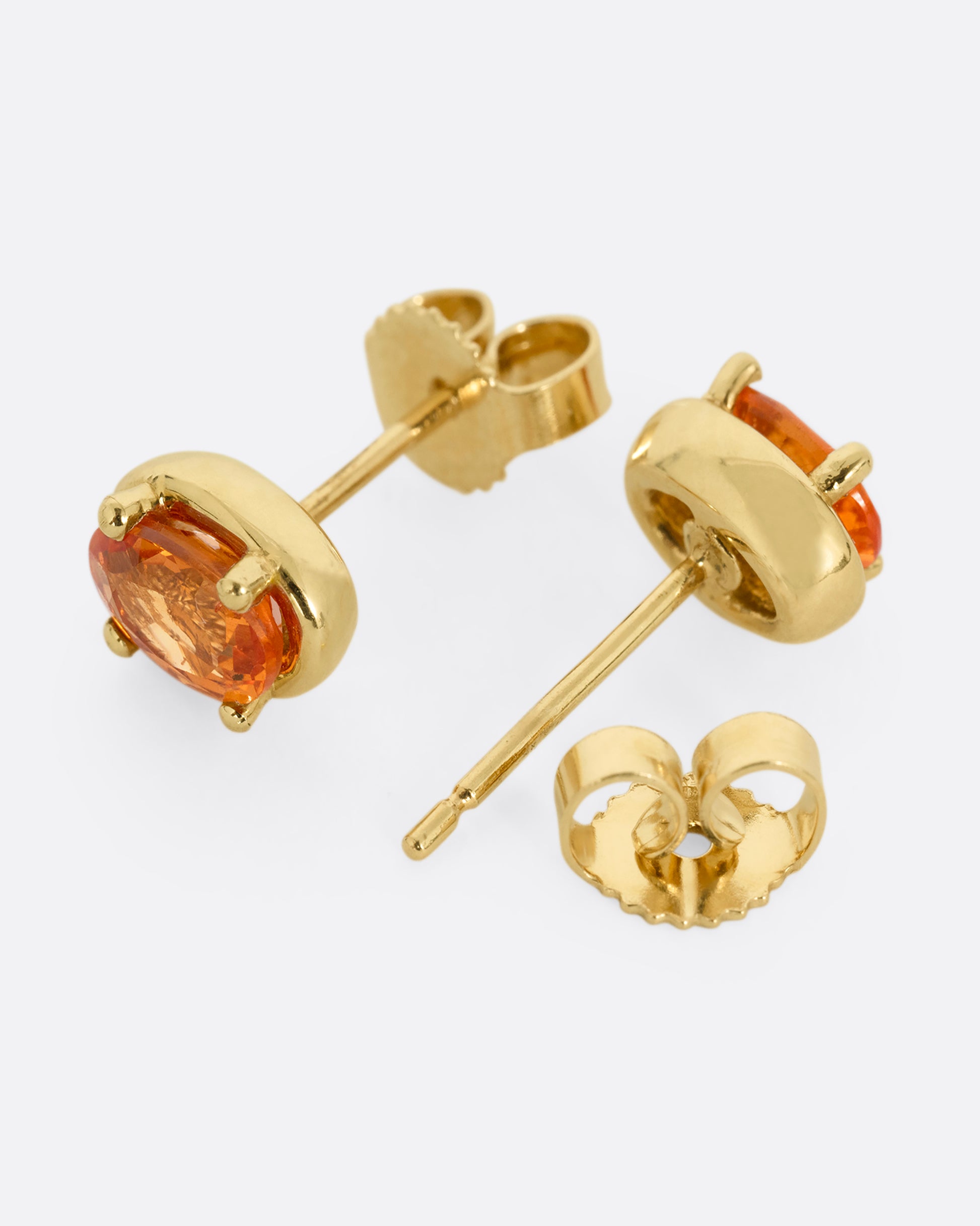 A pair of yellow gold stud earrings with orange sapphires. Shown lying flat from the side; one facing front, the other open facing back.
