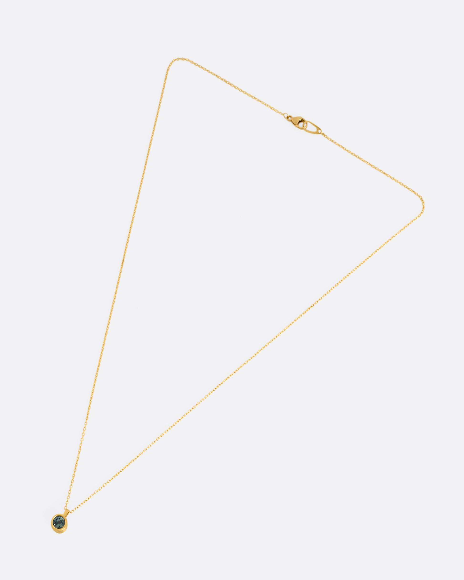 A yellow gold cable chain necklace with a round bezel set faceted blue green sapphire. Shown lying flat clasped from the front.