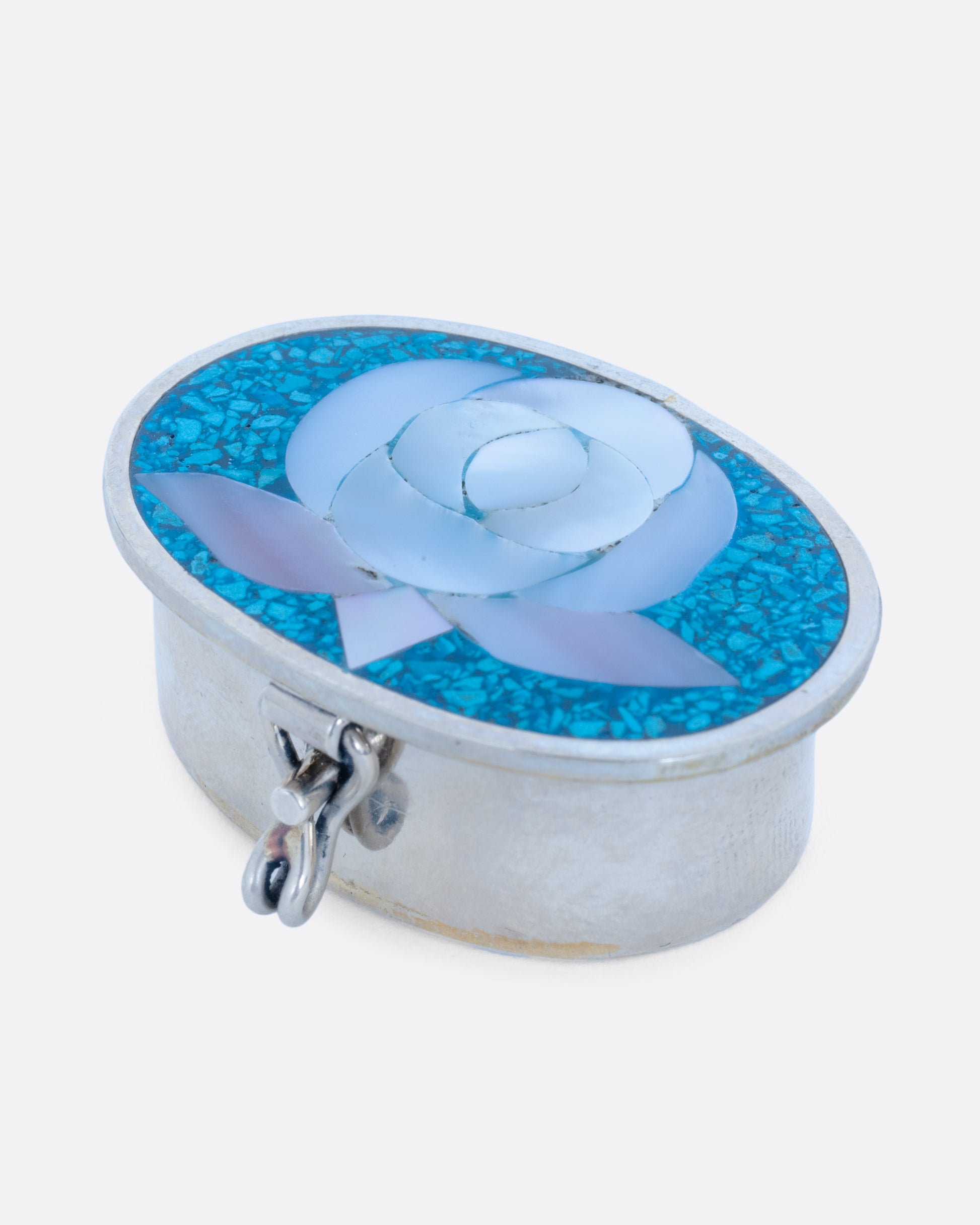 An oval alpaca silver box with a turquoise and mother of pearl flower motif top and closure in front. Shown at an angle from the front.