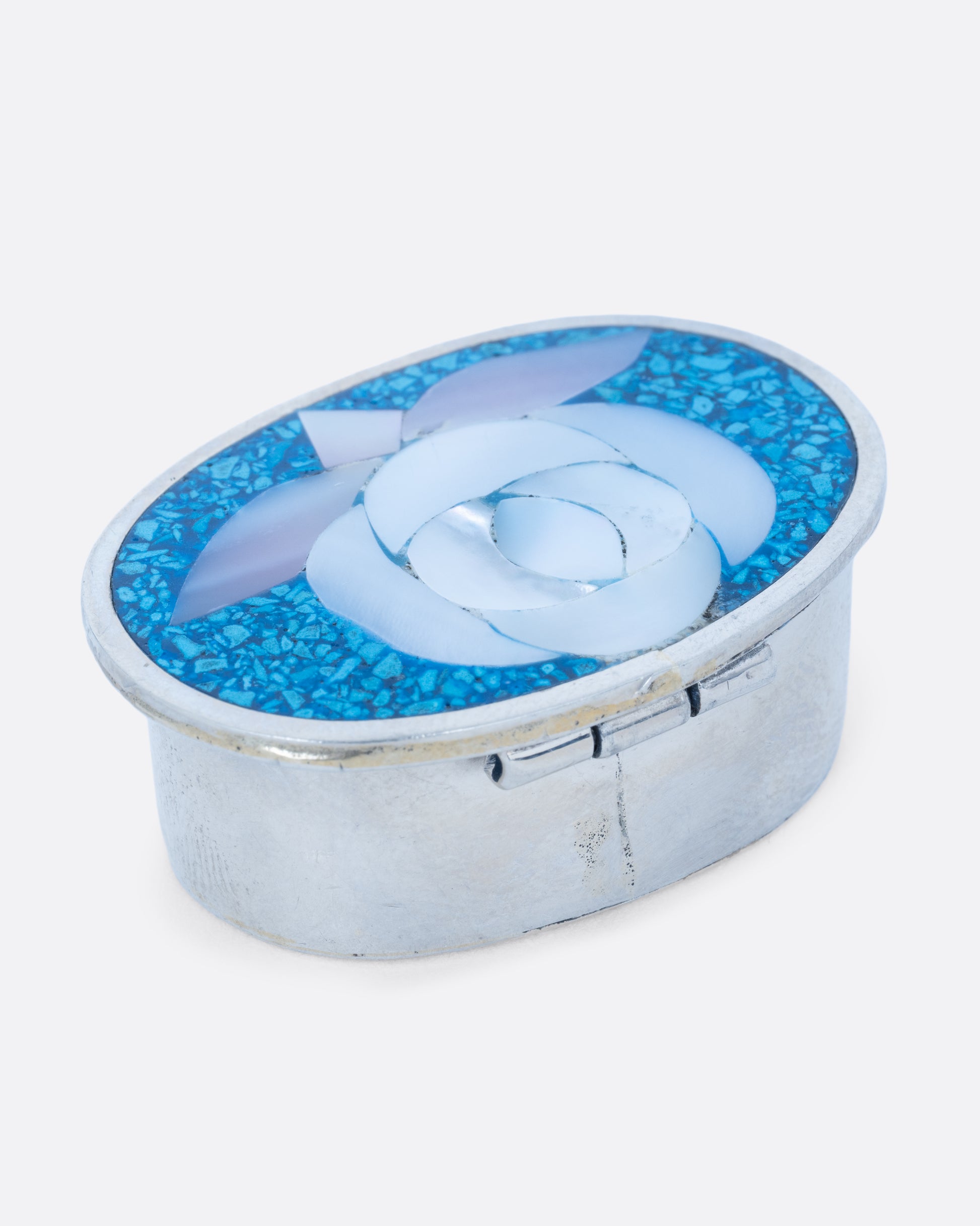 An oval alpaca silver box with a turquoise and mother of pearl flower motif top and closure in front. Shown at an angle from the back.