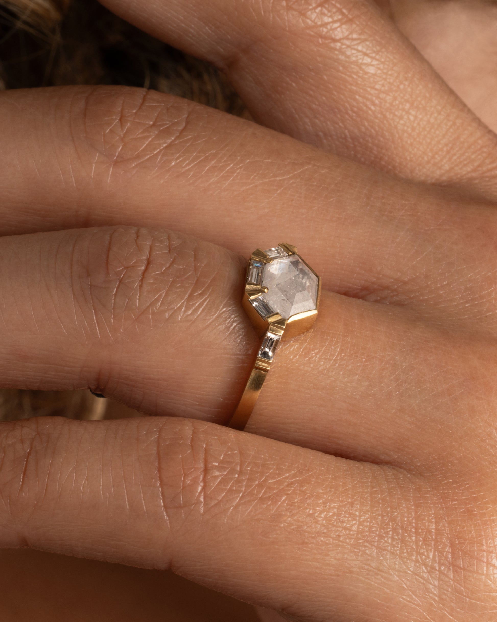 This ring features an east-west set, hexagonal, rose cut gray diamond with a half-halo of baguette diamonds. Shown worn on a finger.