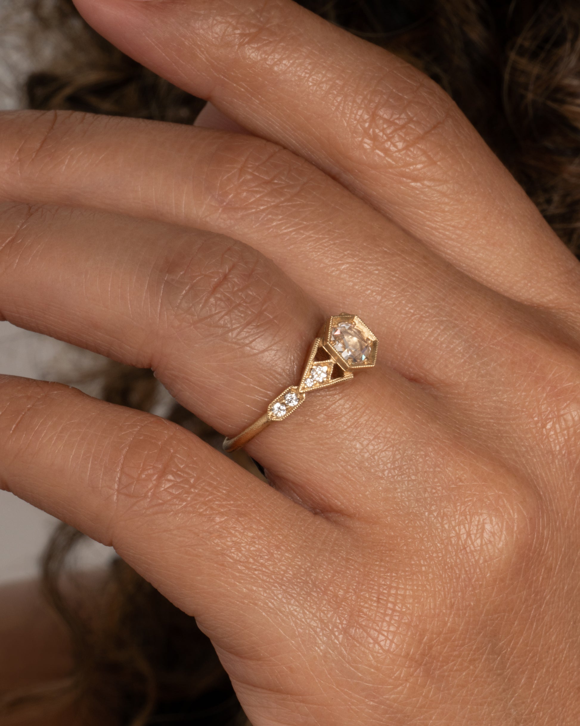 A yellow gold ring with a center diamond and angular side cutouts and diamond accents. Shown from the side on a hand.