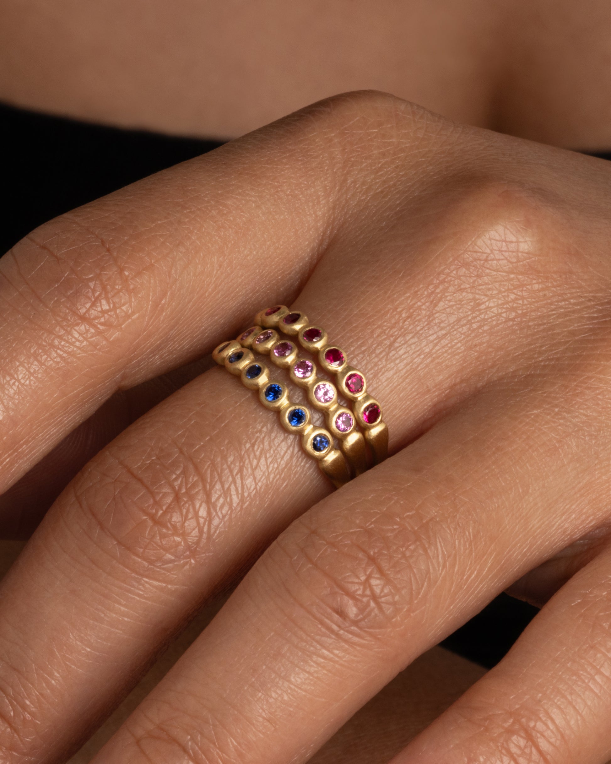 A yellow gold band with a row of pink sapphires. Shown stacked with a ruby and blue sapphire skimmer band worn on a finger.