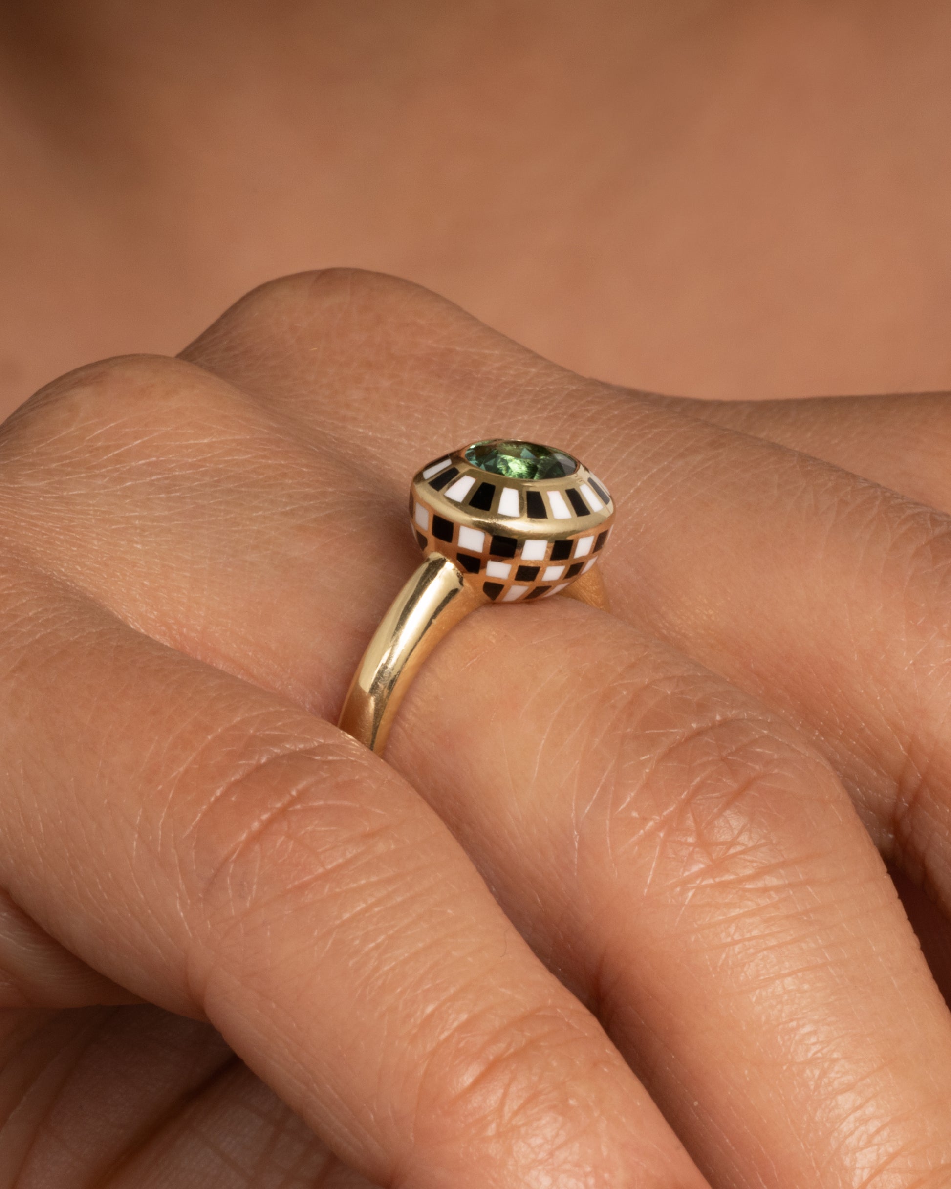A yellow gold ring with an enamel black and white checkerboard pattern framing a green tourmaline. Shown from the front on a hand. 