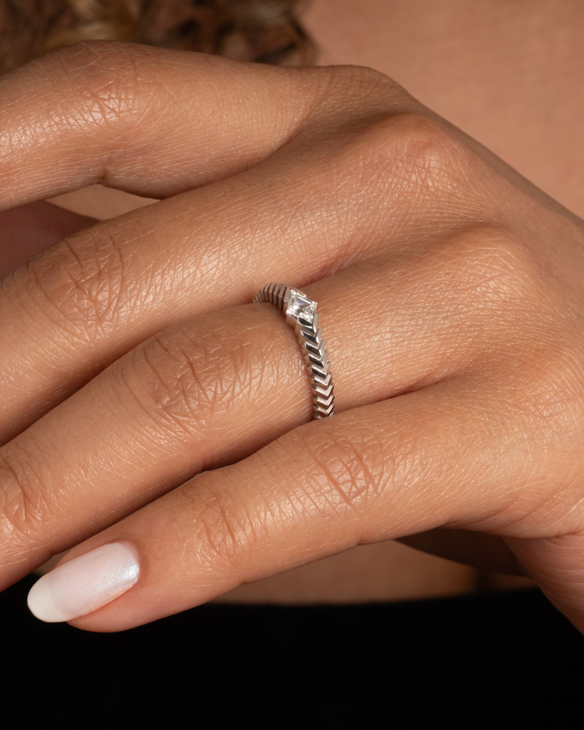 A platinum ring with a 'pleated' motif and lozenge shaped diamond at the center. Shown from the side on a hand.