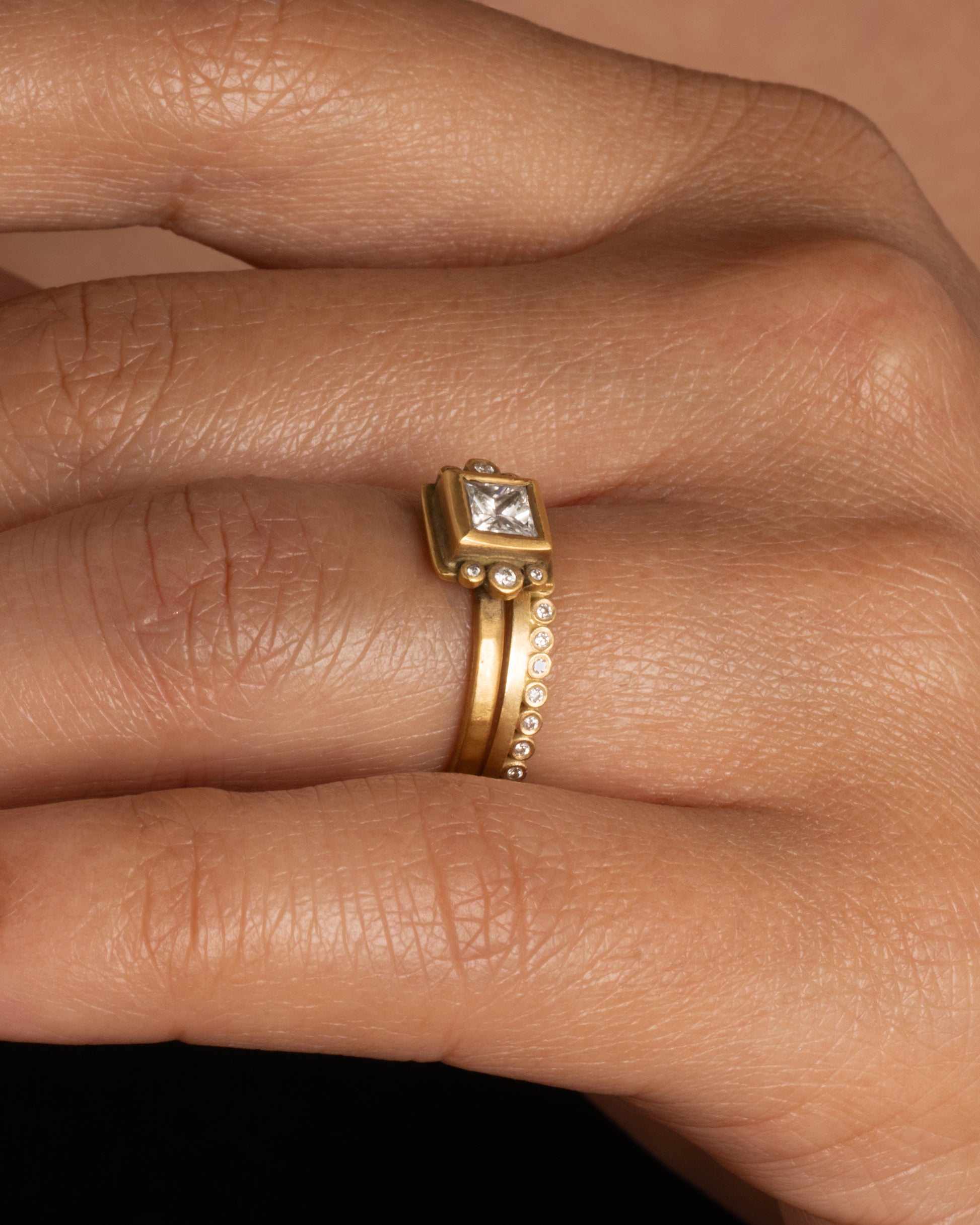 A hand with two rings; a yellow gold ring with bezel set diamond dots encircling one side, and a yellow gold band with a square central diamond and bezel diamond details. Shown from the side.