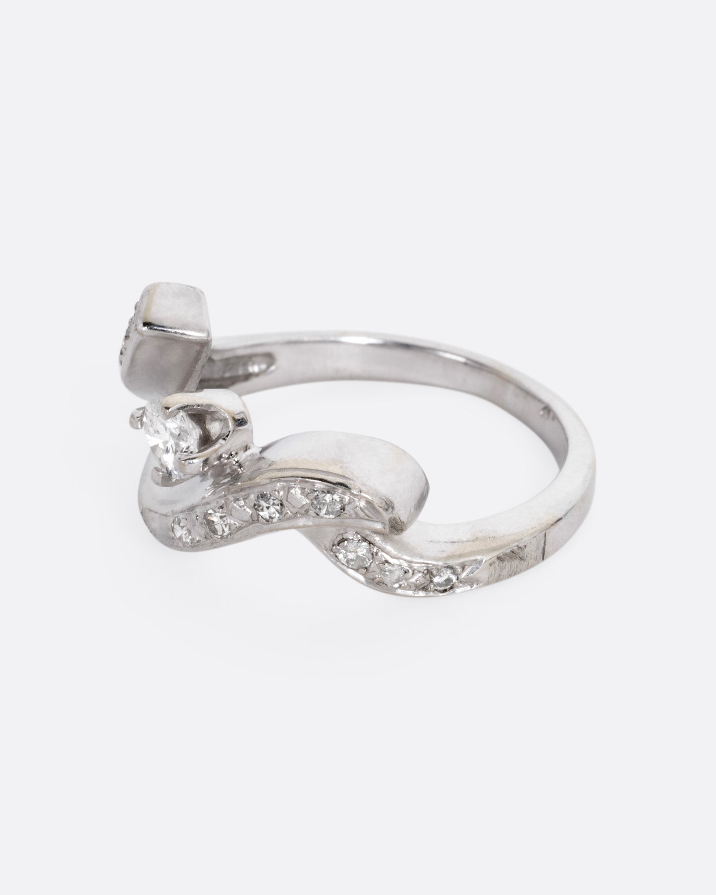 A white gold ring with a curving freeform silhouette, bead set diamonds, and a brilliant cut prong set central diamond, shown from the side.