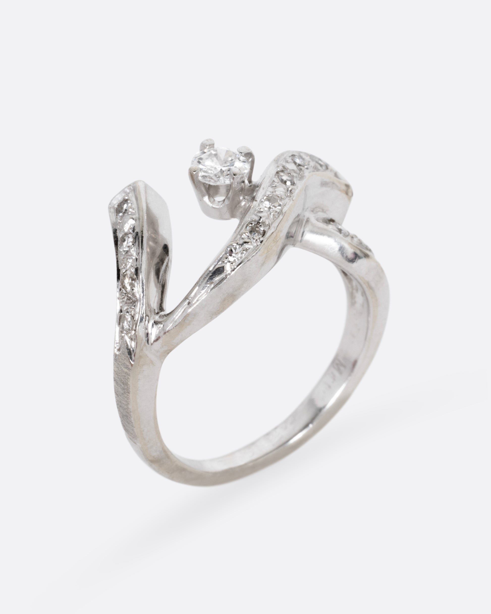 A white gold ring with a curving freeform silhouette, bead set diamonds, and a brilliant cut prong set central diamond, shown standing up from the side.