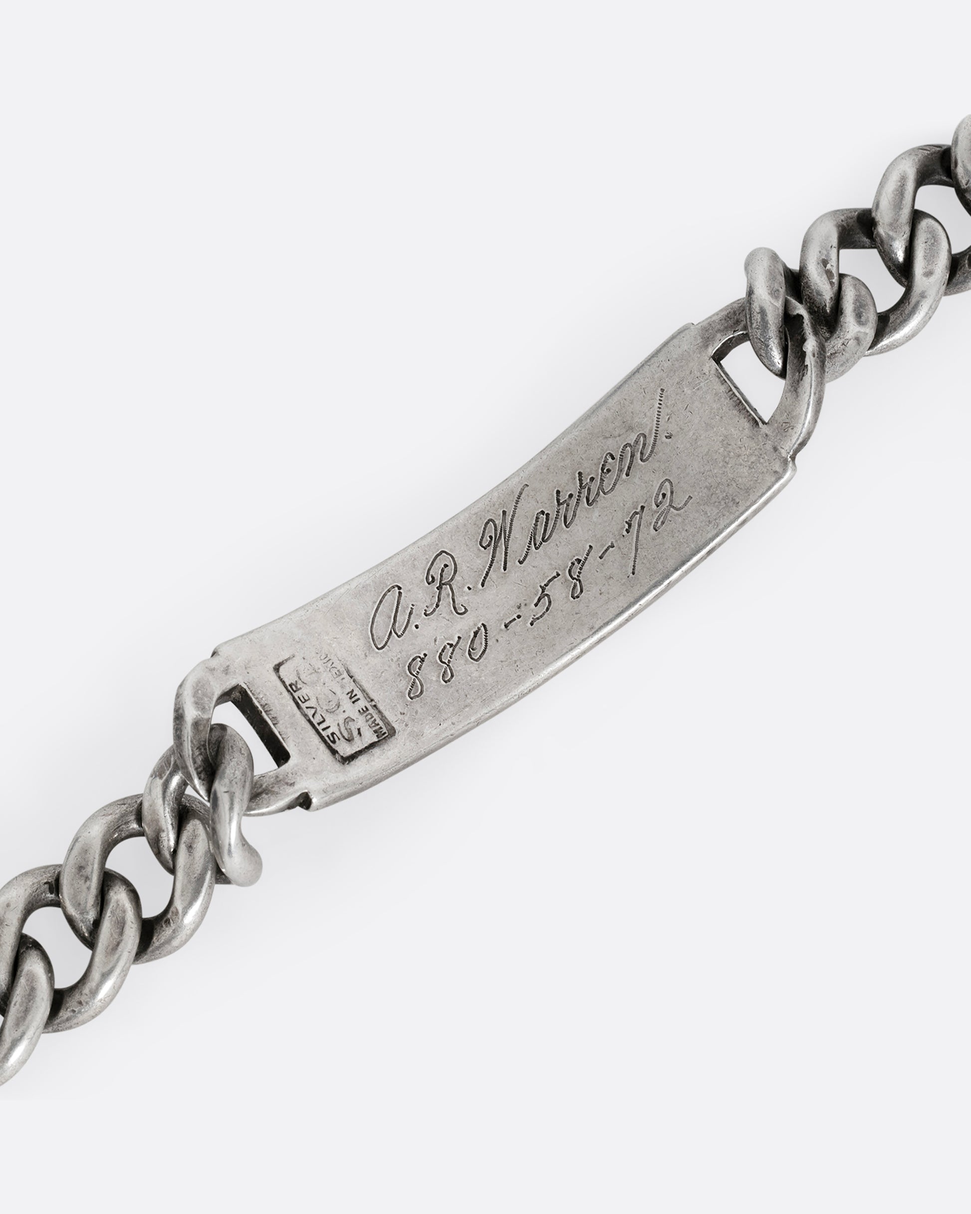 A close up of a silver cable chain ID bracelet with "A.R. Warren" in curling script  engraved on the inside of its name tag. 