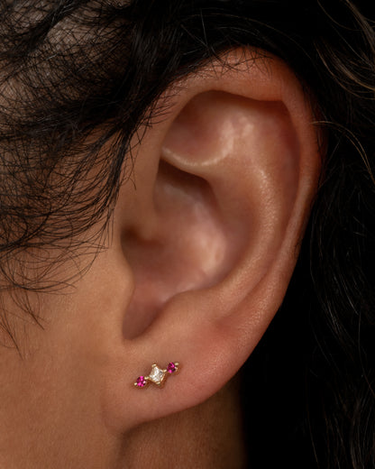 A yellow gold stud earring with a princess cut diamond at the center and two round pink sapphires on either side.  Shown from the front worn in an ear.