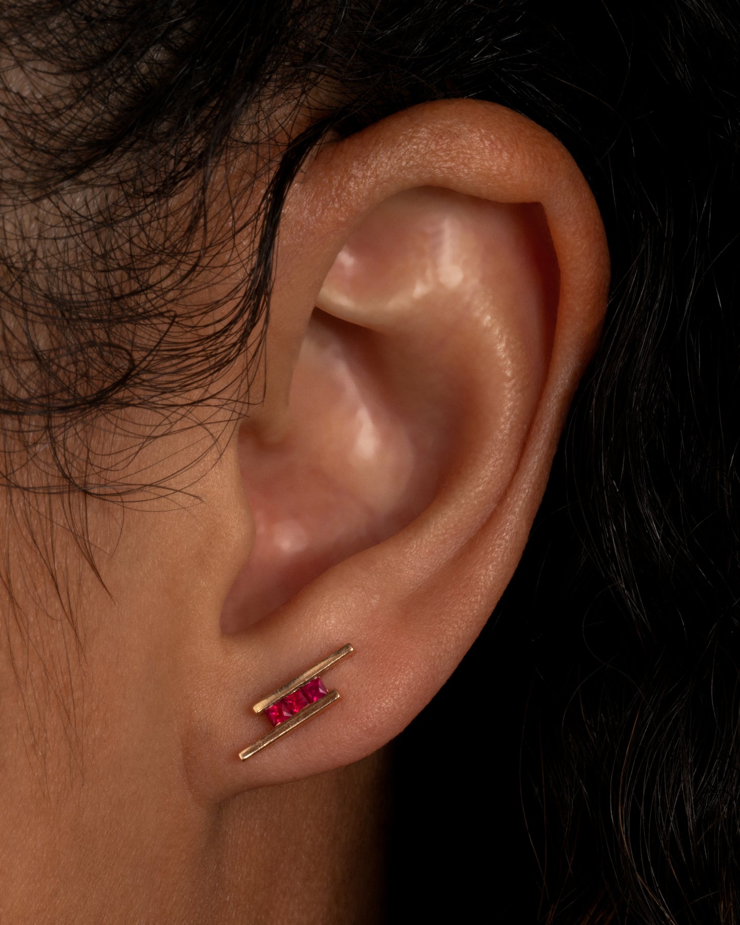 A rose gold stud earring with three rubies between two bars. Shown on an ear.
