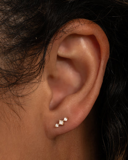 A white gold stud earring with a princess cut diamond at the center and two round diamonds on either side. View from the front on a person.