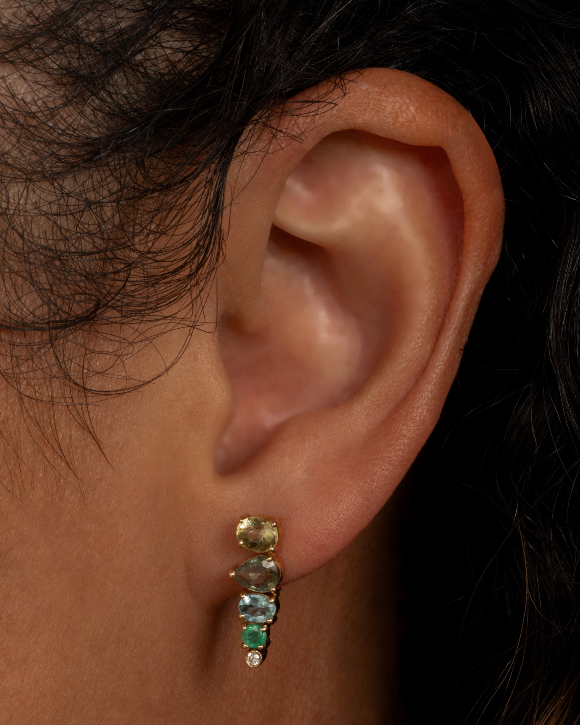 A yellow gold stud earring with a mosaic of emerald, sapphire, tourmalines and diamonds. Shown worn in an ear.
