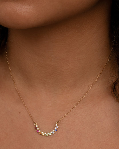 A yellow gold cable chain necklace with a crescent shaped pendant, studded with multicolored gemstones, shown on a person.