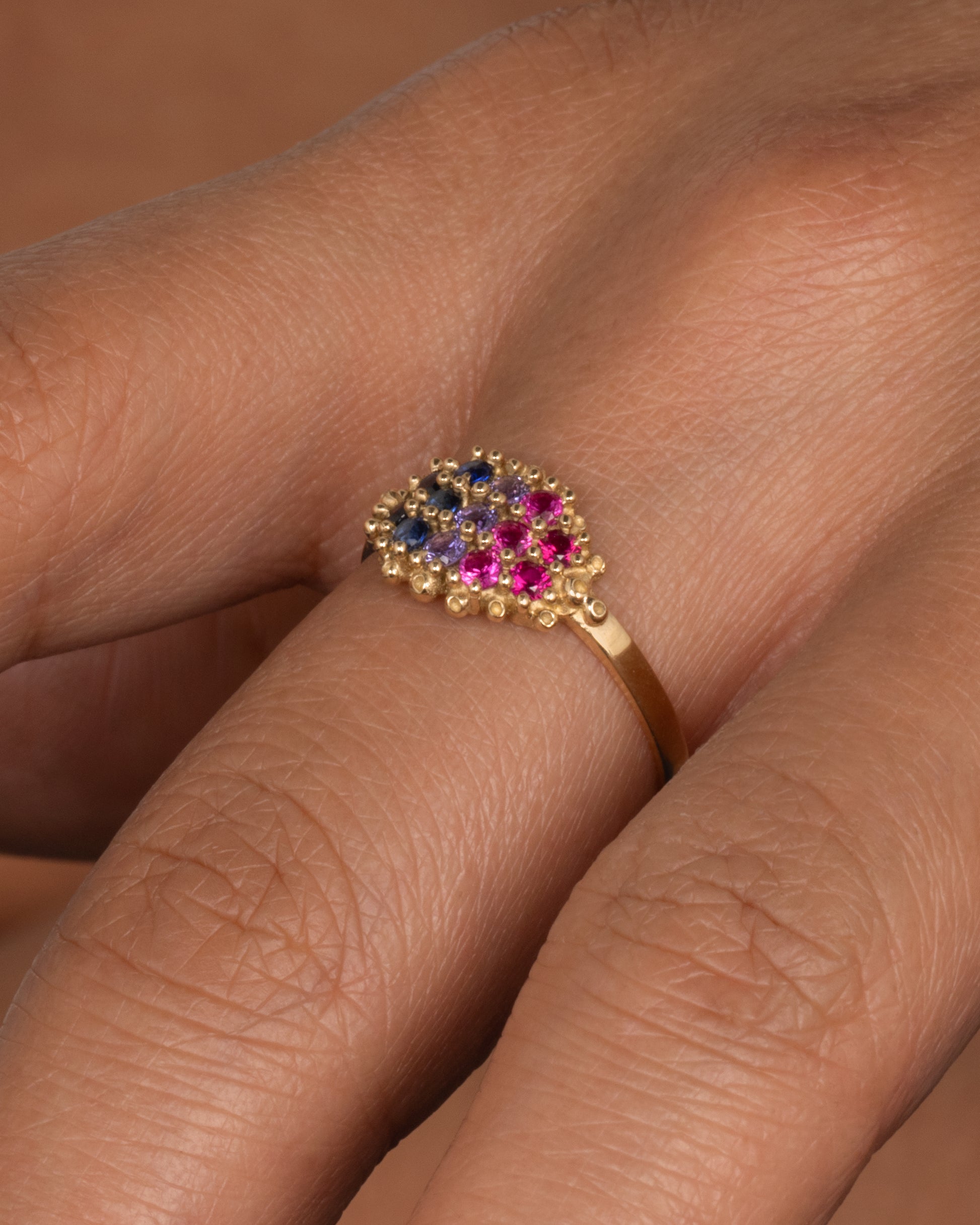A yellow gold signet style ring whose face is lined with ombré pink to blue sapphires in textured prong settings. Shown worn on hand.