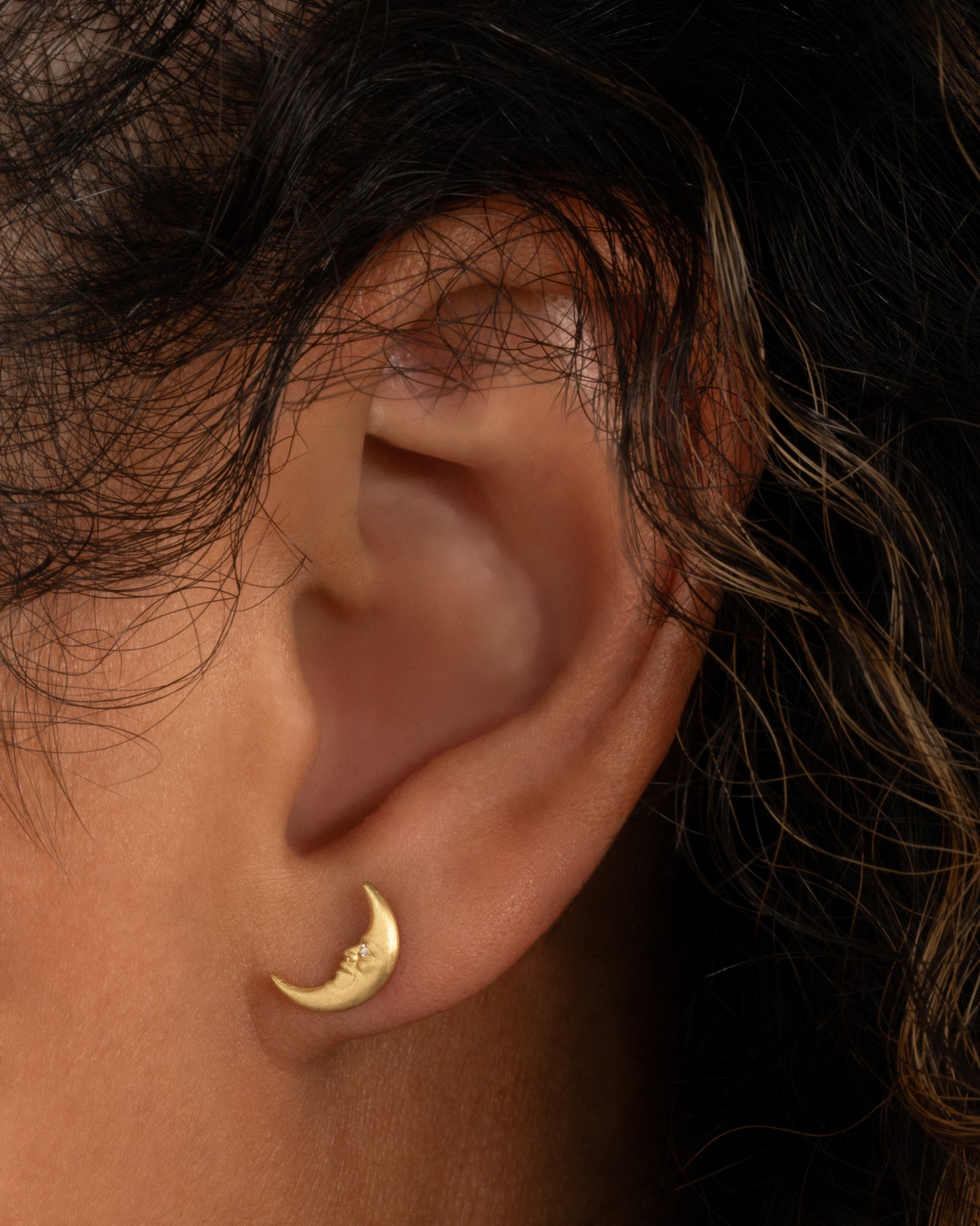 A yellow gold crescent moon stud earring with a face and a diamond eye. Shown worn in an ear.