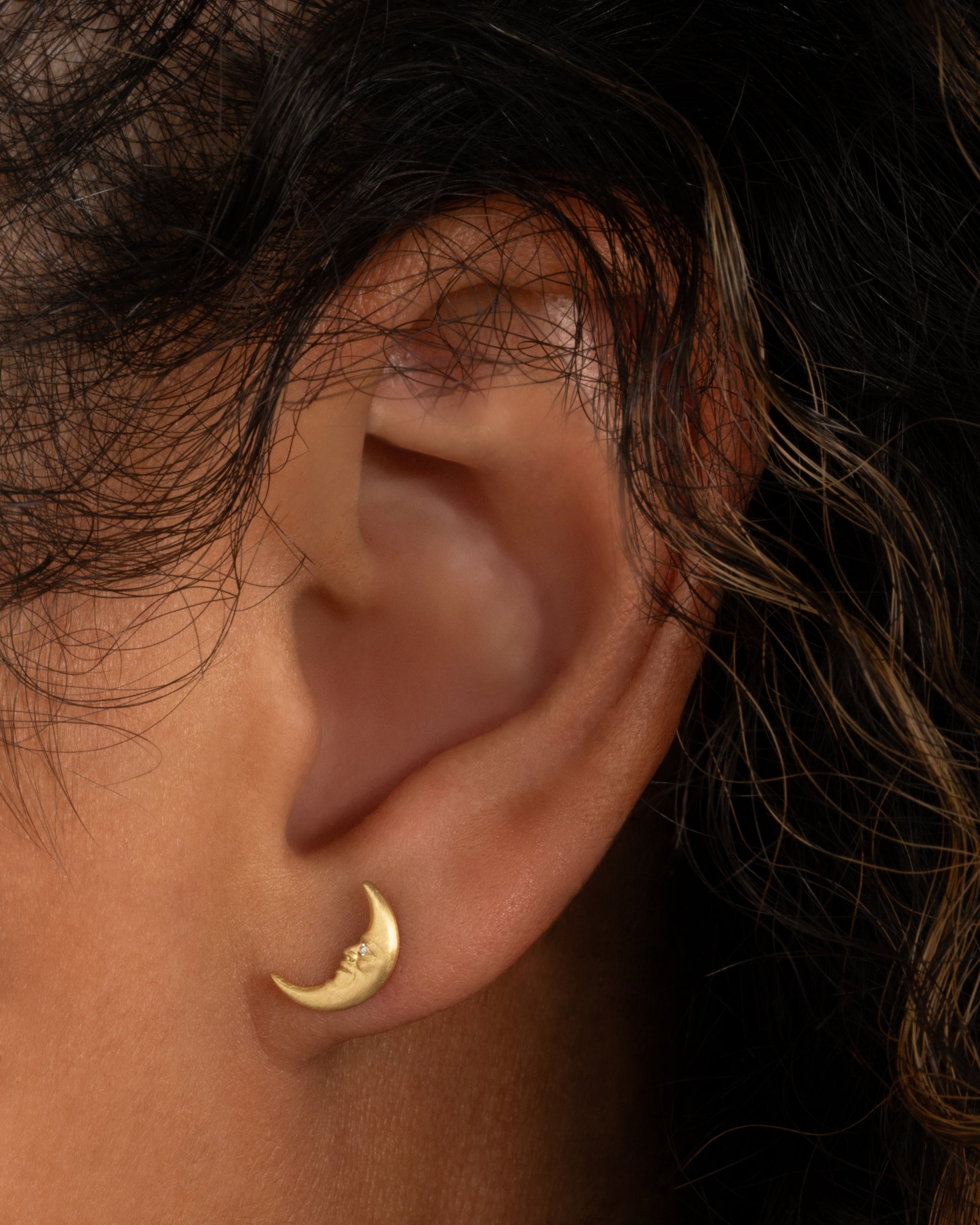 A yellow gold crescent moon stud earring with a face and a diamond eye. Shown worn in an ear.