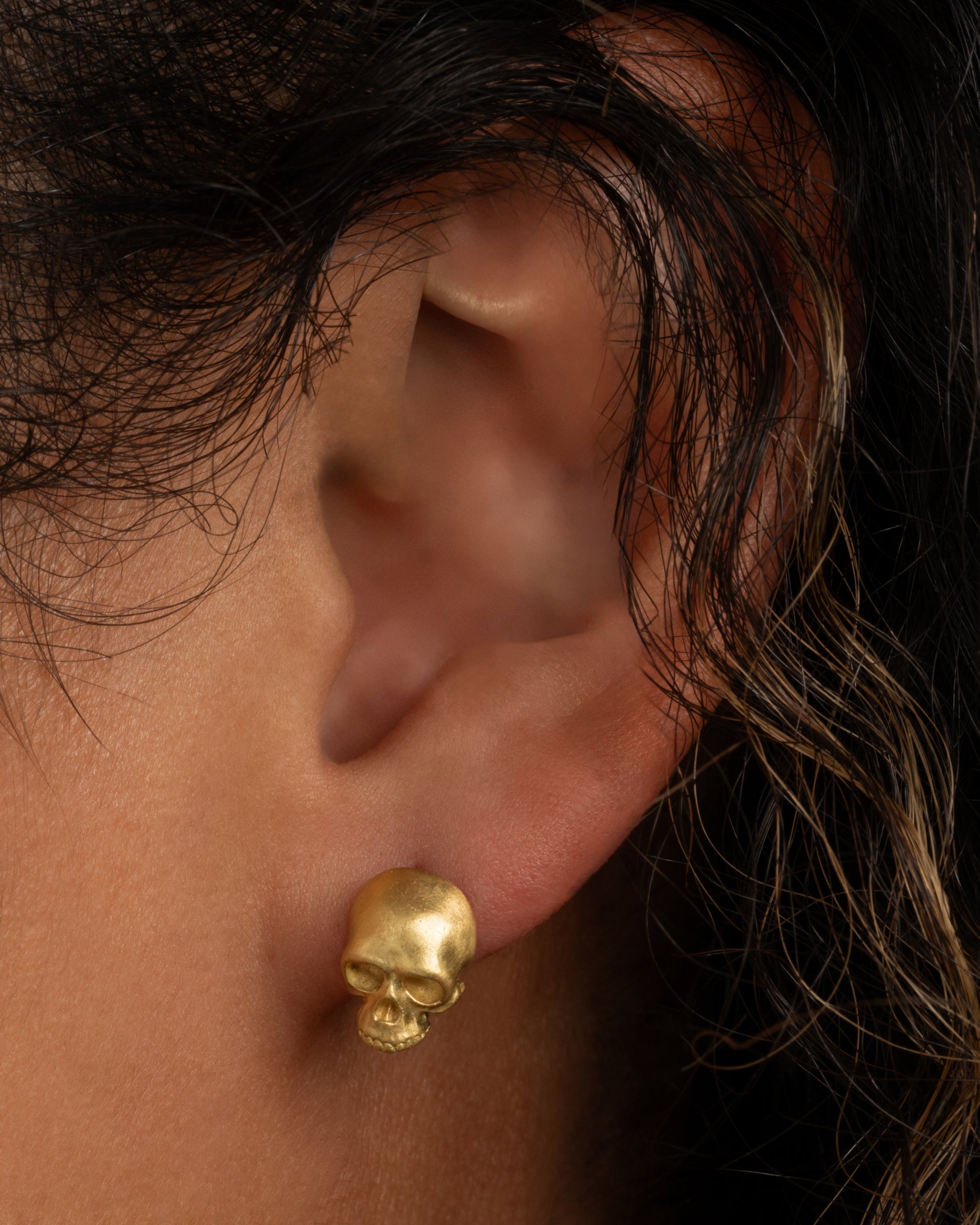 A solid yellow gold skull stud earring. Shown worn in an ear.