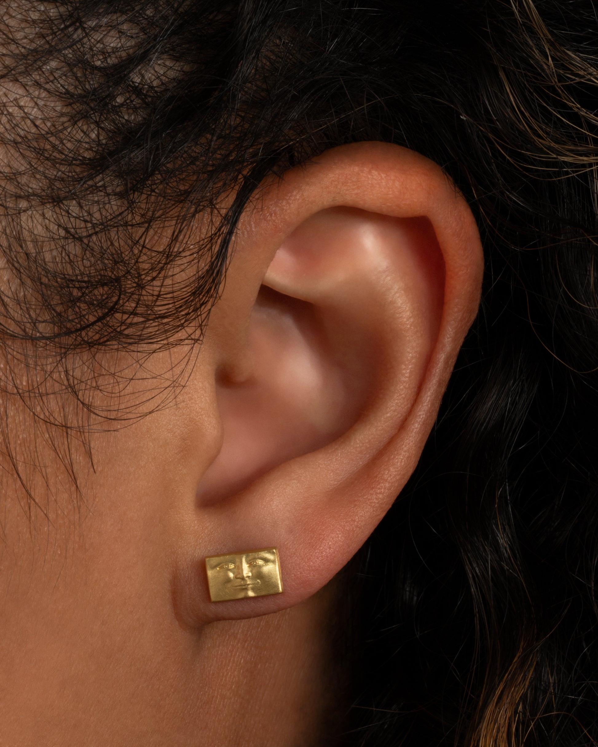 A yellow gold rectangular brick stud earring with a face. Shown worn in an ear.
