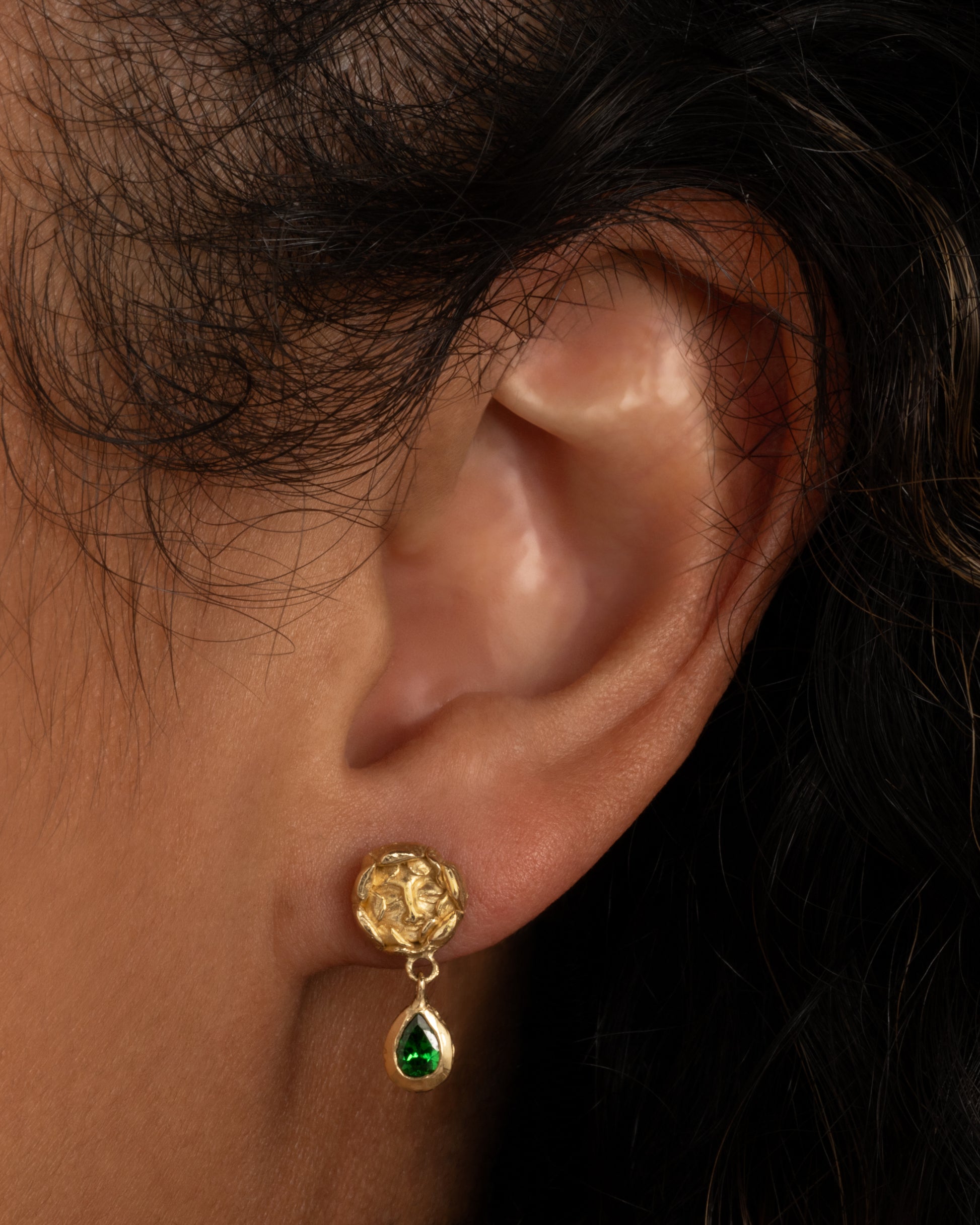 A pair of yellow gold studback earrings with faces at the center of rosebuds and a pear shaped tsavorite dangle. Shown worn in an ear.
