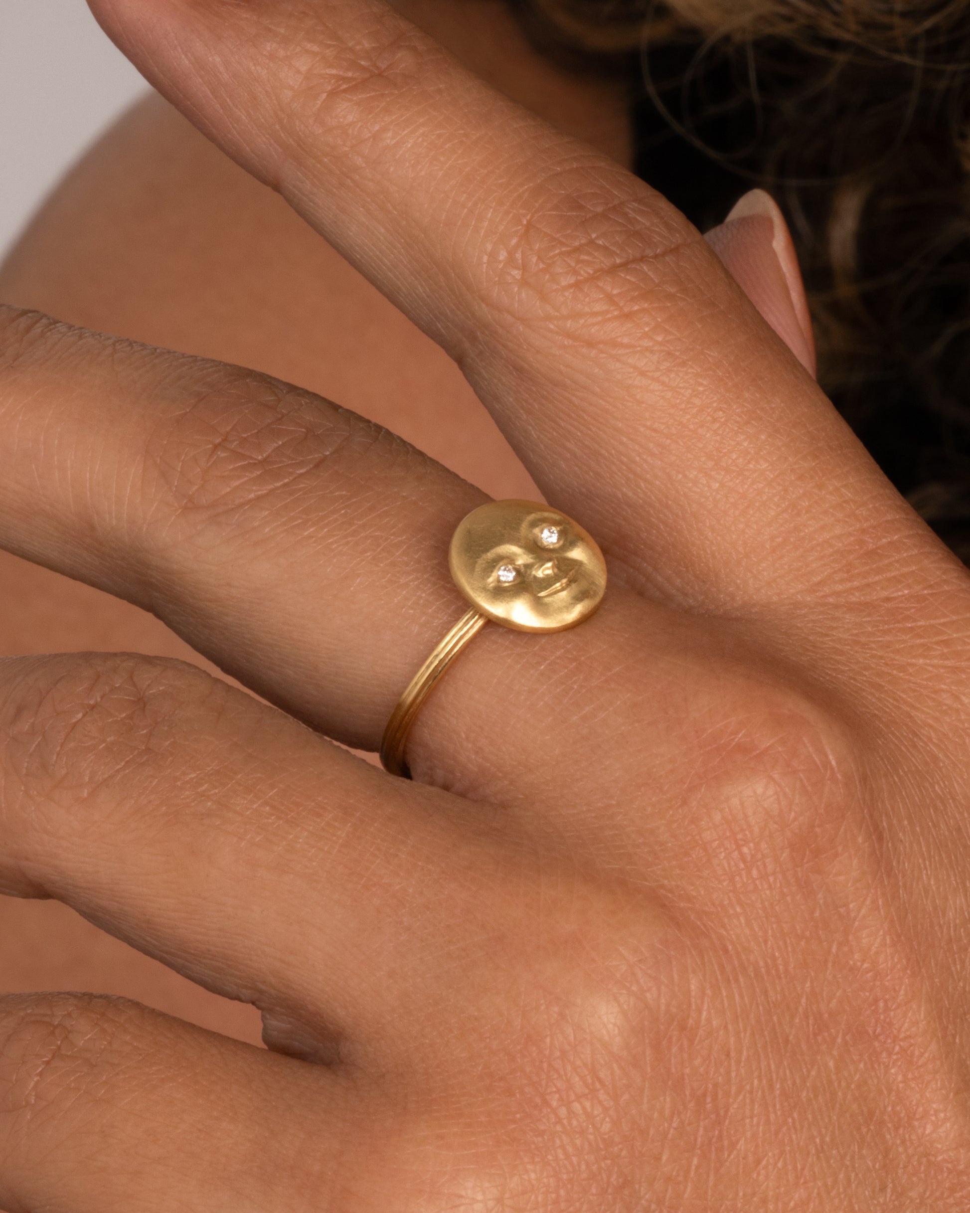 A yellow gold ring with a round moon face with diamond eyes. Shown worn on hand.