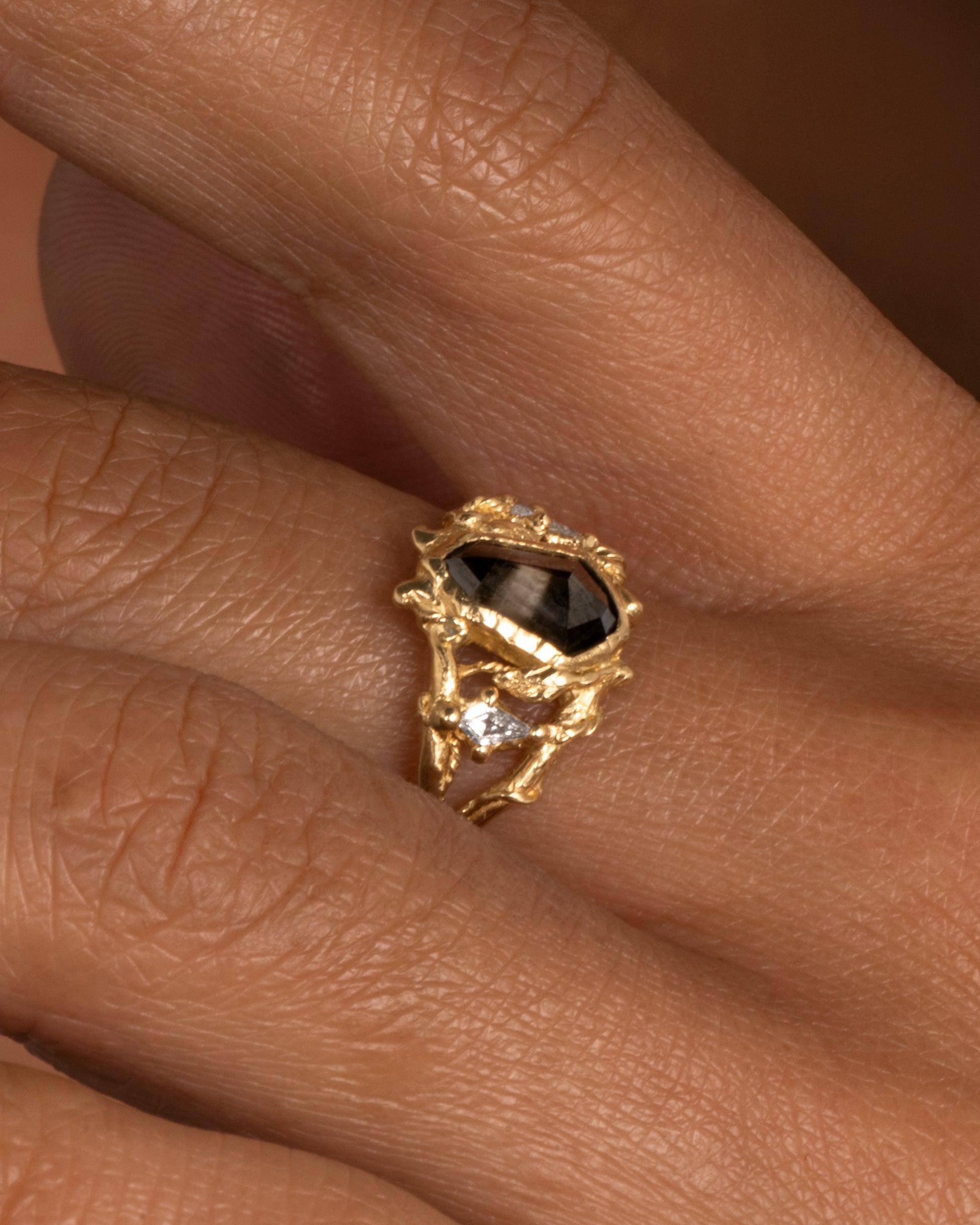 A yellow gold ring with an organic vine motif and a shield double cut black diamond at the center with white kite sapphires on either side. Shown worn on hand.