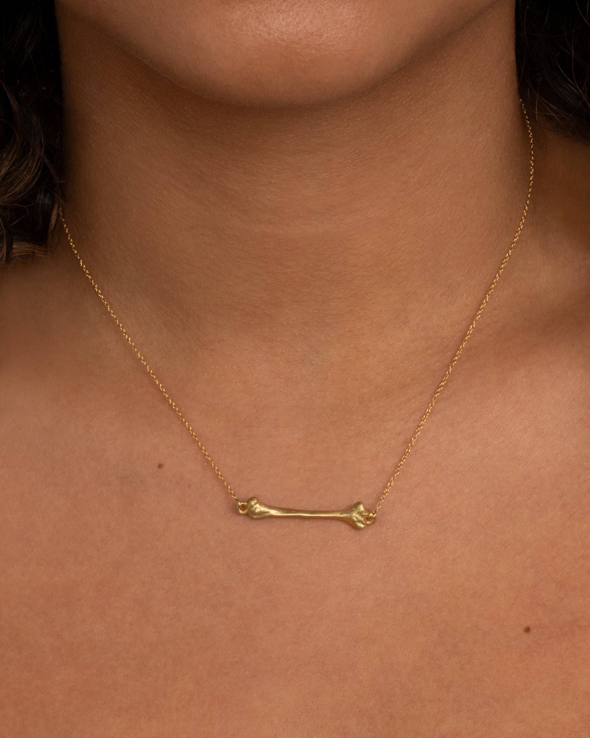 A yellow gold cable chain necklace with a bone charm horizontally suspended from the center. Shown worn around the neck.