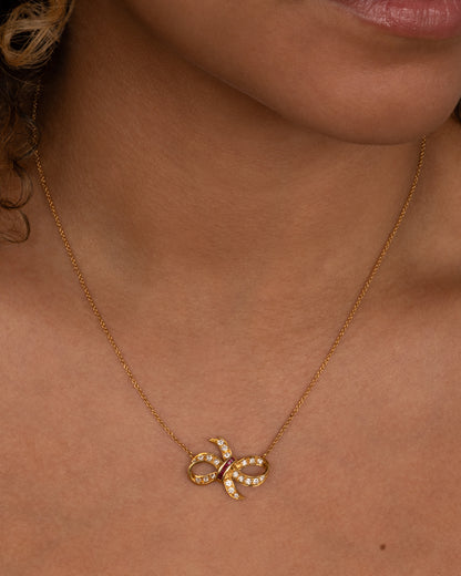 A yellow gold cable chain necklace with a diamond and ruby studded bow at the center. Shown from the front on a person.