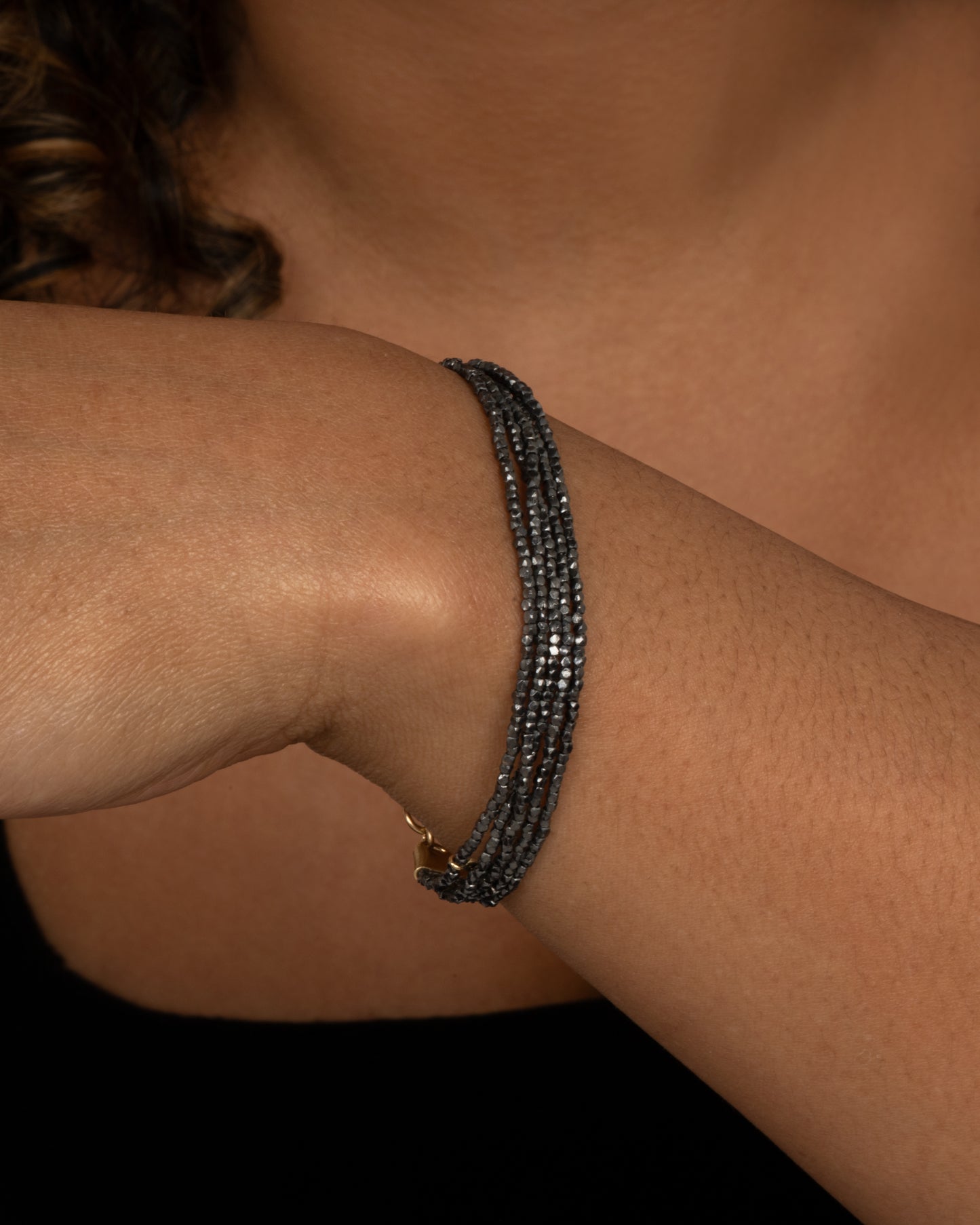 A bracelet with five strands of oxidized silver beads converging at  yellow gold capped ends with a lobster clasp. Shown from the side on a person.