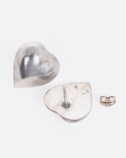 A pair of silver heart shaped stud earrings. One ear shown from the front, the other is open from the back.