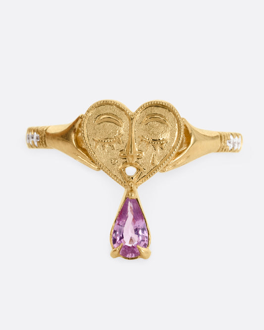 A yellow gold ring with a heart shaped face and diamond studded arms. A pear shaped pink tourmaline sits at the base. Shown from the front.