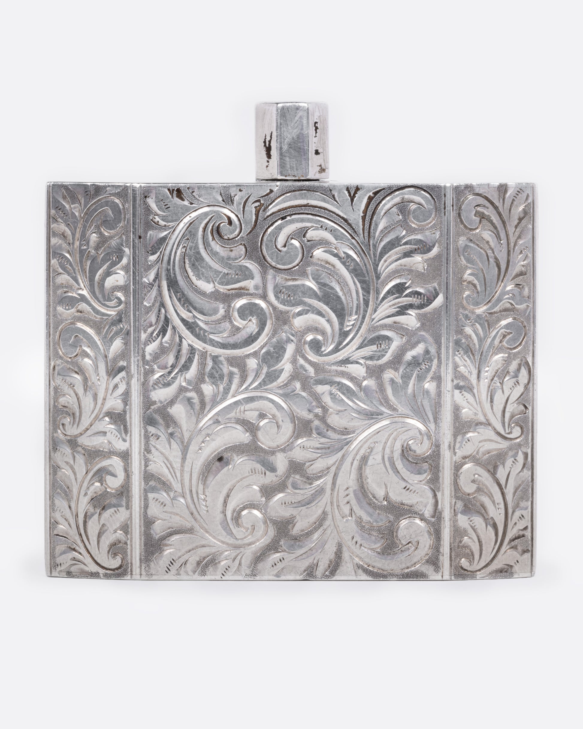A silver perfume bottle with winding botanical etchings and a slim square silhouette, shown from the front.